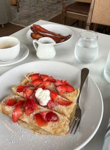 A plate shows a pastry topped with sliced strawberries and whipped cream, dusted with powdered sugar, with a fork. Warm coffee, water, and bacon in background, sunny table setting.