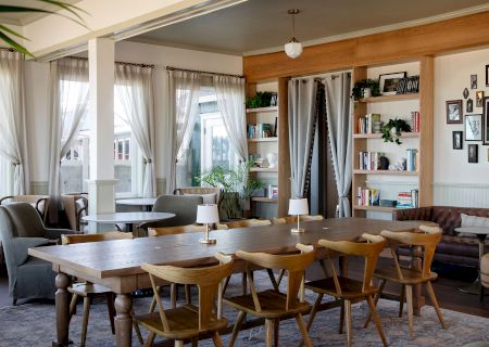 A bright, cozy dining space with a long wooden table and mismatched chairs, bookshelves, plants, and a soft, airy vibe with draped windows.