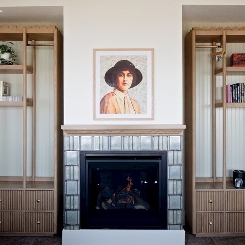 A cozy living room with a tiled fireplace, two wooden bookshelves, and a portrait of a woman in a hat centered above the mantle.