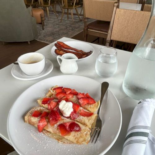 A plate of waffles topped with fresh strawberries and cream, with a side of bacon, plus a coffee, water, and a glass bottle in a bright cafe.