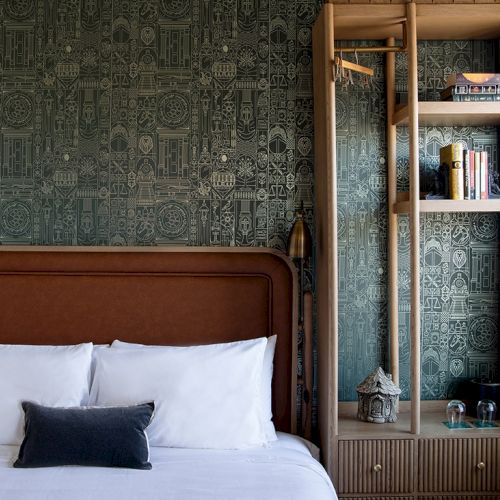 A cozy bedroom features a bed with white linens and a dark pillow, a vintage patterned green wallpaper, and an open wooden shelving unit with decor.