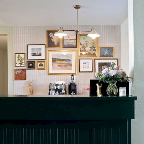 A cozy coffee bar with a dark counter, wall gallery of framed art photos, and a small bouquet on the right, warm lighting overhead.