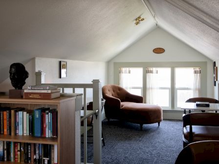 A bright loft living room with bookshelves, a brown armchair, dark carpet, and large windows with curtains.