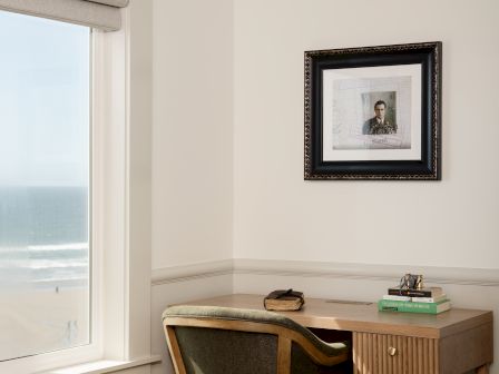 A small home office corner with a wooden desk and chair, books, a framed portrait, and a window with roller blinds.