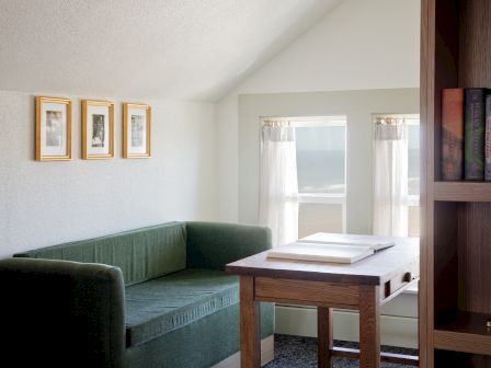 A cozy corner with a green sofa, wooden table, bookshelves, framed pictures, and bright window curtains.