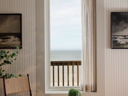 A cozy room with a large window, sheer curtains, ocean view, and a small table with plants and books.