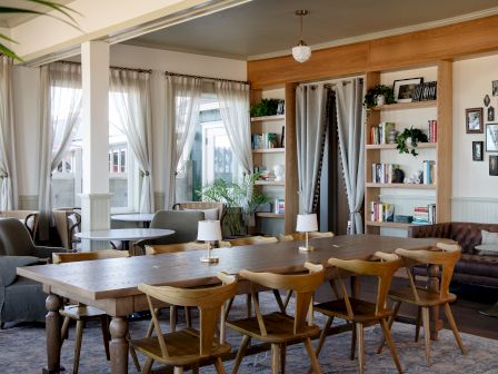 Bright dining/living room with a wooden table, chairs, grey curtains, and built-in shelves full of books and decor.