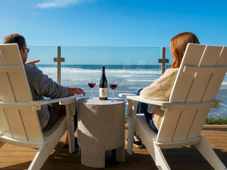 Two people relax in white beach chairs with wine bottles and glasses on a terrace, facing the ocean and waves on a sunny day.