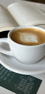 A latte and an open book on a white table, with a green menu card nearby, creating a cozy reading moment.