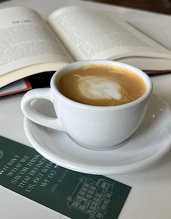 A latte and an open book on a white table, with a green menu card nearby, creating a cozy reading moment.