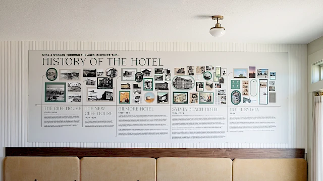 A white wall with a large &ldquo;History of the Hotel&rdquo; timeline poster above a tan cushioned bench in a bright lobby, soft natural light.