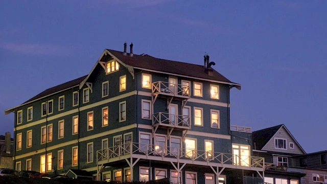Green multi-story house at dusk with lit windows and balconies, neighboring building, calm blue sky, warm indoor lights.