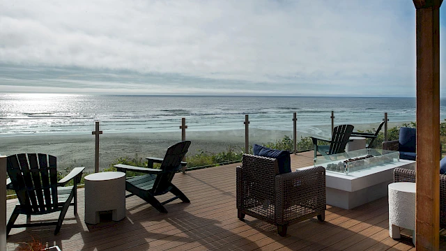 A sunny beachfront deck with chairs, a table, and a hot tub, overlooking calm ocean waves under a partly cloudy sky.