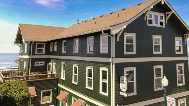 A dark green multi-story house with white trim, brown roof, numerous windows, and a clear blue sky above.