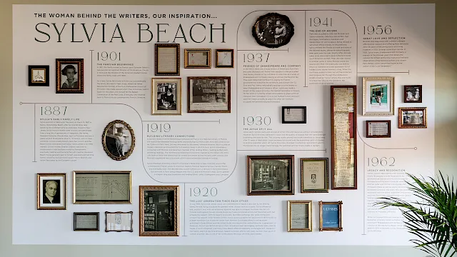 A caf&eacute; wall shows a framed timeline poster for Sylvia Beach, with chairs and a pendant light above.