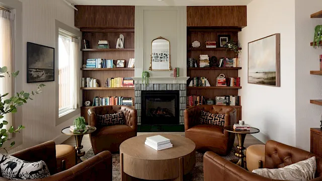A cozy living room with a central fireplace flanked by built-in bookshelves, brown leather chairs, a round coffee table, and sunlit windows.