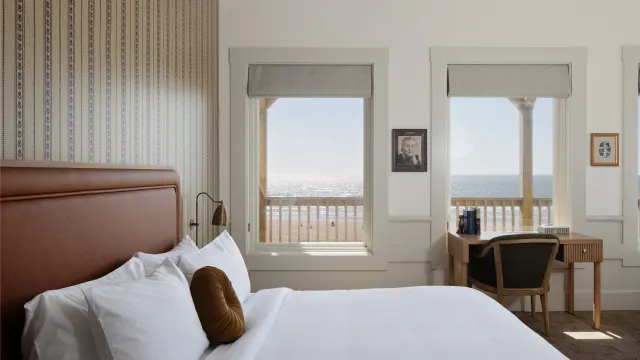A bright seaside hotel room with a large bed, desk and chair, and two windows showing the ocean.
