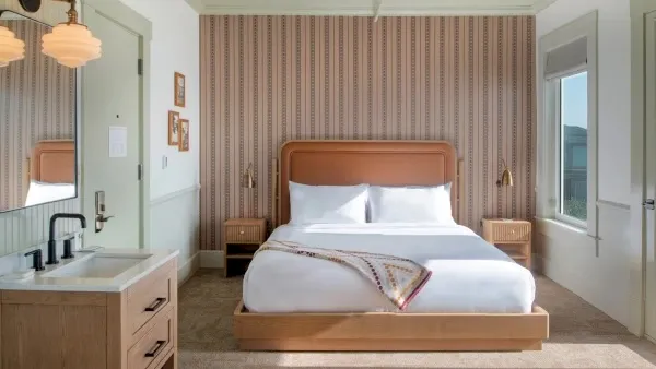 A cozy hotel room with a wooden bed, striped accent wall, bedside tables, a small vanity, and soft natural light from windows on both sides.