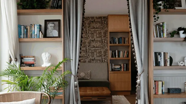 A cozy living room with built‑in bookshelves flanking glass doors, a plant in the foreground, and a doorway opening to a wood‑paneled study with a patterned wall, ceiling beams, and a warm, bookish vibe.