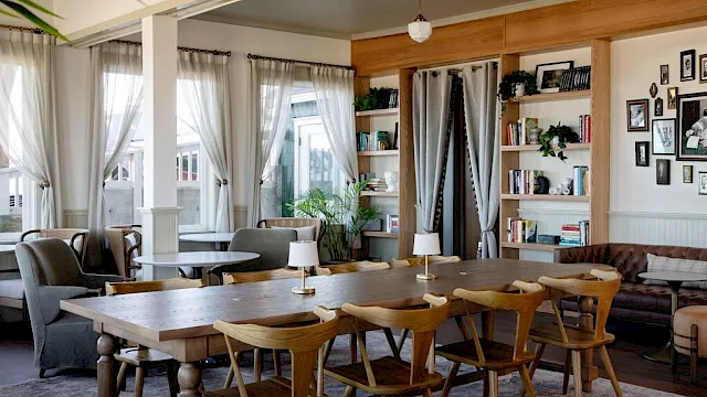 A bright, cozy living space with a long wooden dining table, mid-century chairs, bookshelves, lounge areas, plants, and soft natural light.