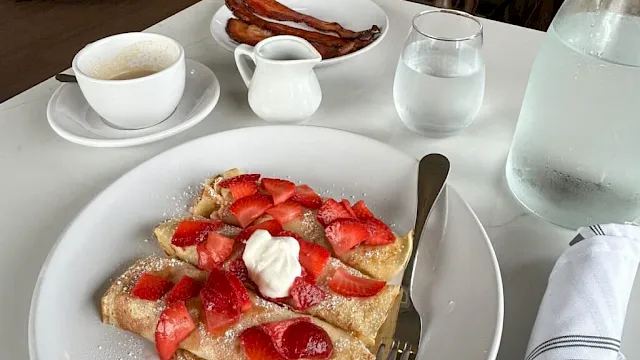 A plate with a nutty pastry topped with fresh strawberries and whipped cream, beside a cup of coffee, water, and crispy bacon in the background.