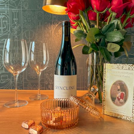 A bottle of wine, two champagne flutes, a rose bouquet, chocolates, and a &ldquo;William Shakespeare Sonnets & Poems&rdquo; book on a table.