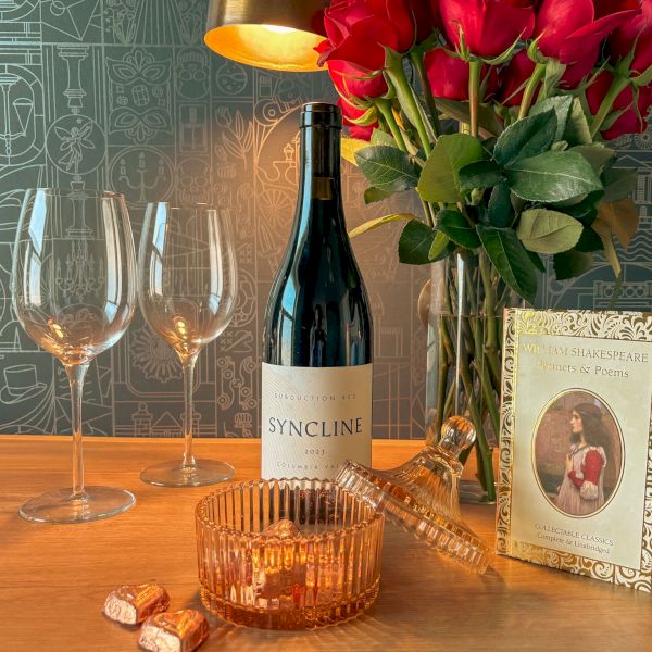 A bottle of wine, two champagne flutes, a rose bouquet, chocolates, and a &ldquo;William Shakespeare Sonnets & Poems&rdquo; book on a table.