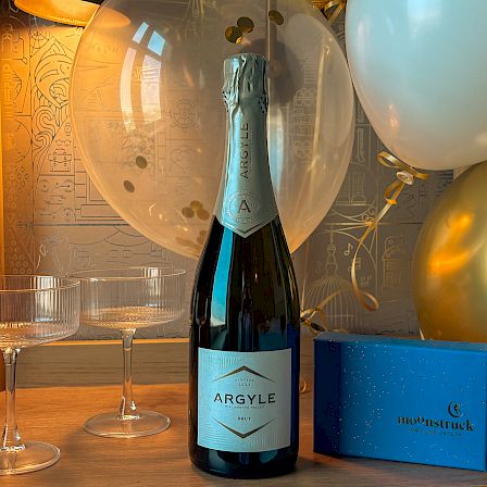 A champagne bottle labeled Argyle Brut with two empty wine glasses, lit by warm lights and set against balloon d&eacute;cor.