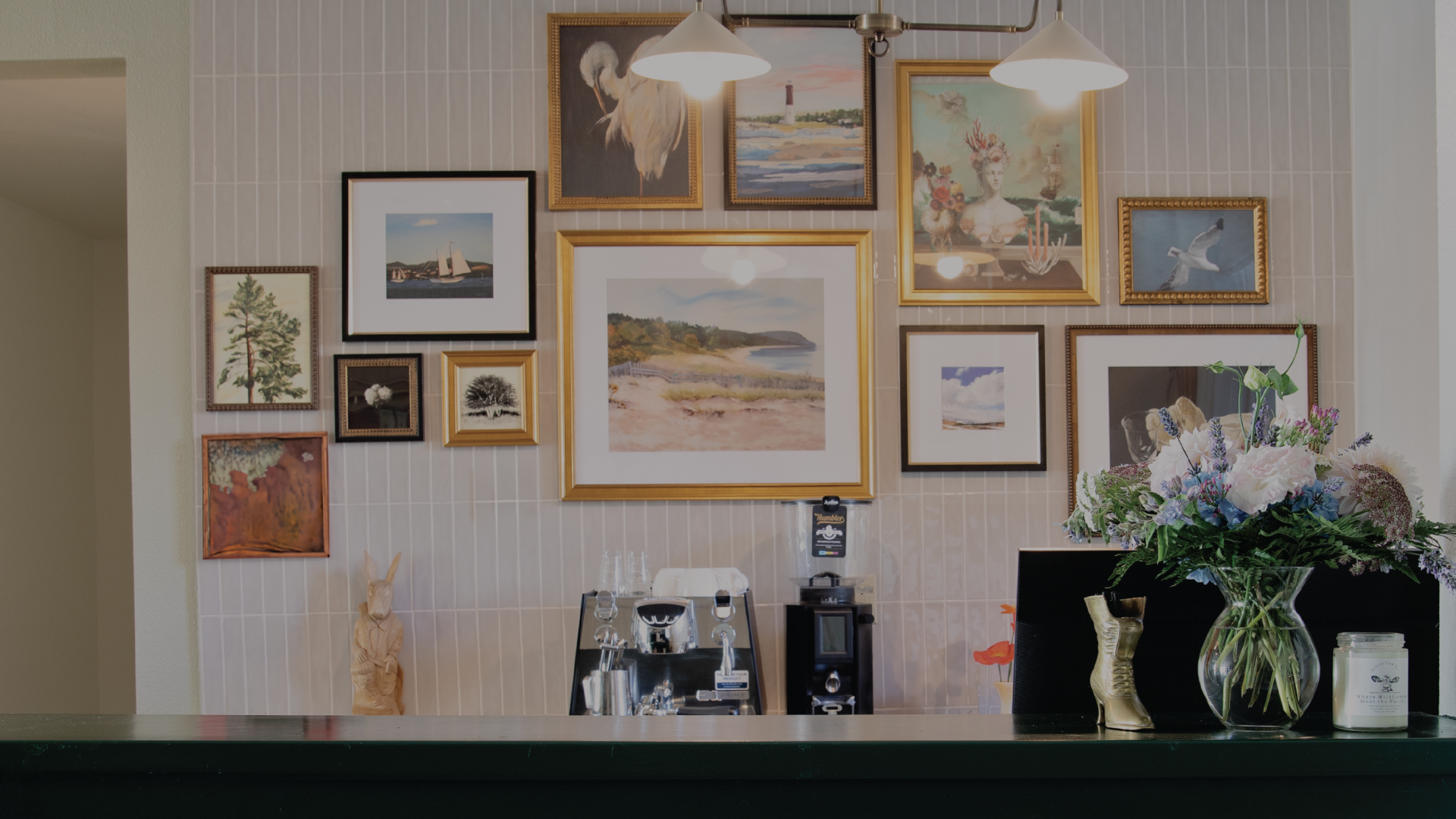A cozy caf&eacute; counter with framed art on tiled walls, hanging lamps, coffee machines, and fresh flowers in vases.