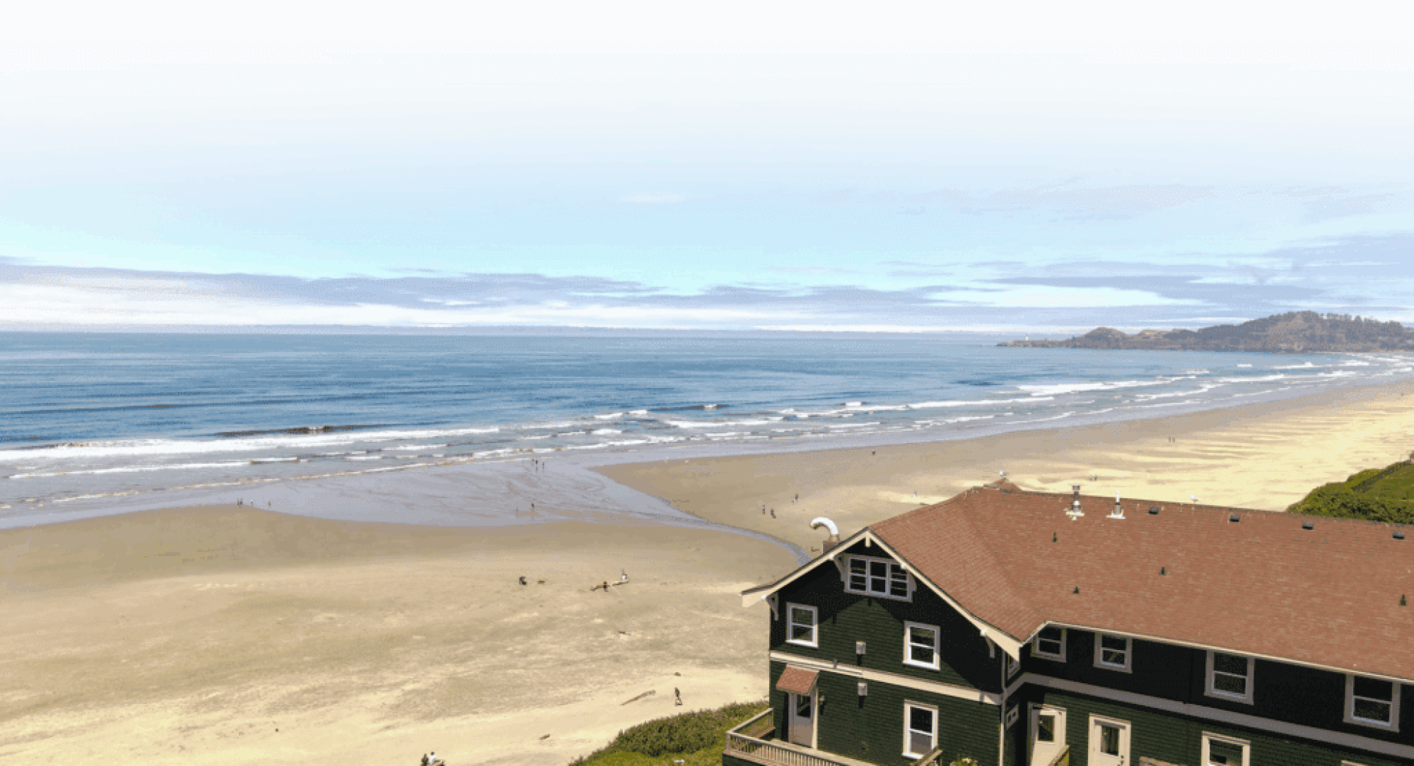 A seaside view with sandy beach, gentle waves, distant hills, and a house overlooking the shoreline.