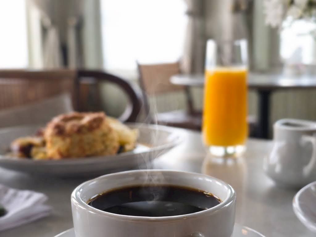 A caf&eacute; table with a steaming cup of black coffee, a glass of orange juice, and pastries in the background, softly blurred.