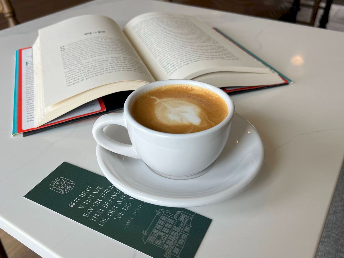 A cup of hot coffee sits beside an open book on a table, with another green book nearby.