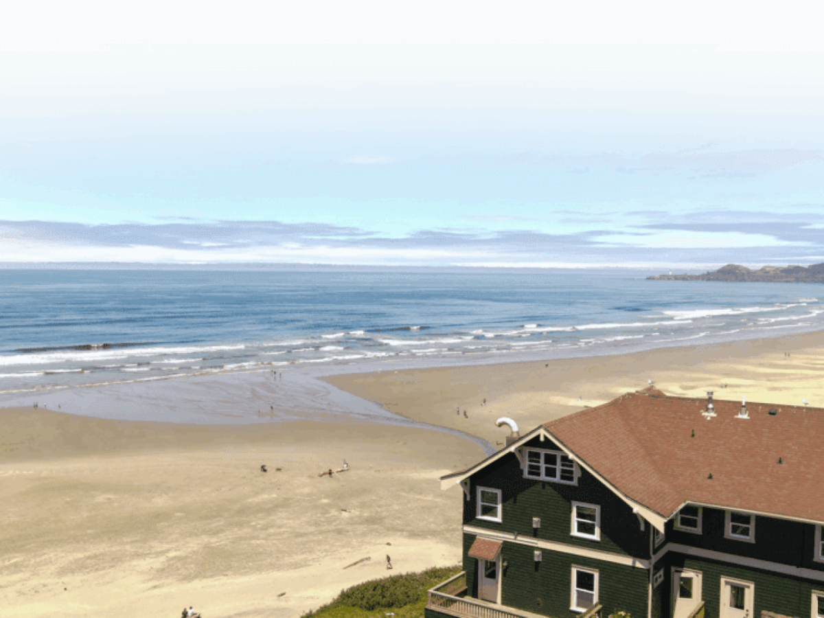 A wide sandy beach with ocean waves under cloudy skies, viewed from a nearby brick-and-green building overlooking the shore.