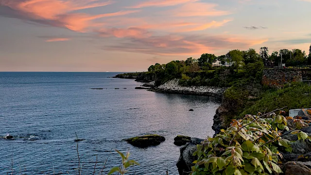 Sunset over a rocky shoreline with calm sea, lush greenery, and distant houses by the coast.