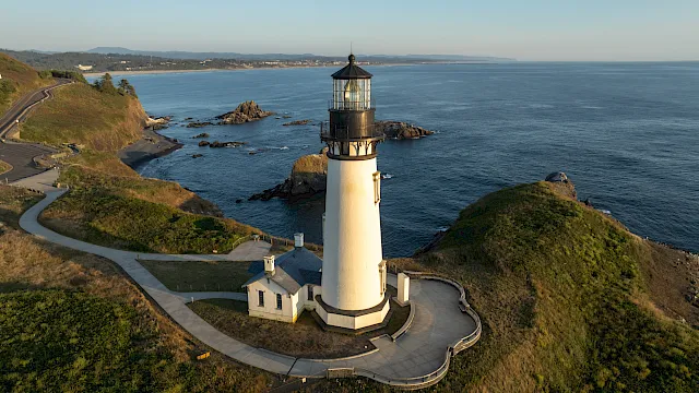 A tall lighthouse with a glass lantern room overlooks a rocky coastline and winding paths along the blue ocean.